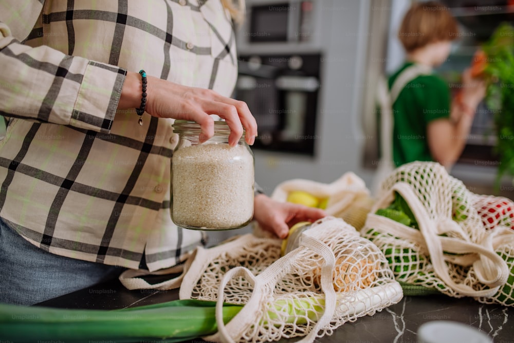 Una madre desempacando alimentos locales en envases de residuo cero de la bolsa con la ayuda de su hija en la cocina de su casa.