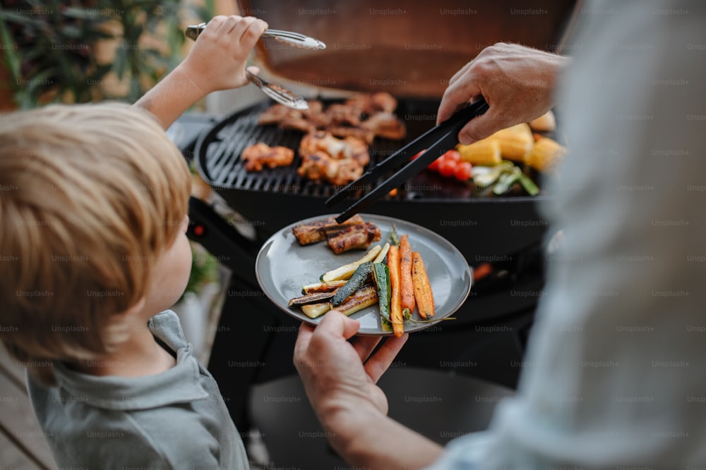 An unrecognizable father with little son grilling ribs and vegetable on grill during family summer garden party, close-up