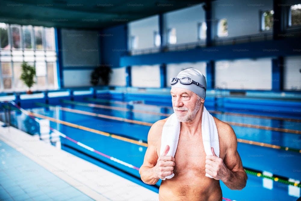 Uomo anziano in piedi vicino alla piscina coperta, braccia sui fianchi. Pensionato attivo che ama lo sport.