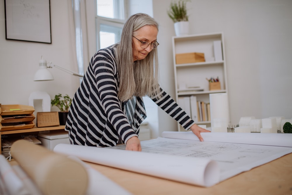 Une femme architecte senior avec une maquette de maisons regardant des plans dans un bureau.