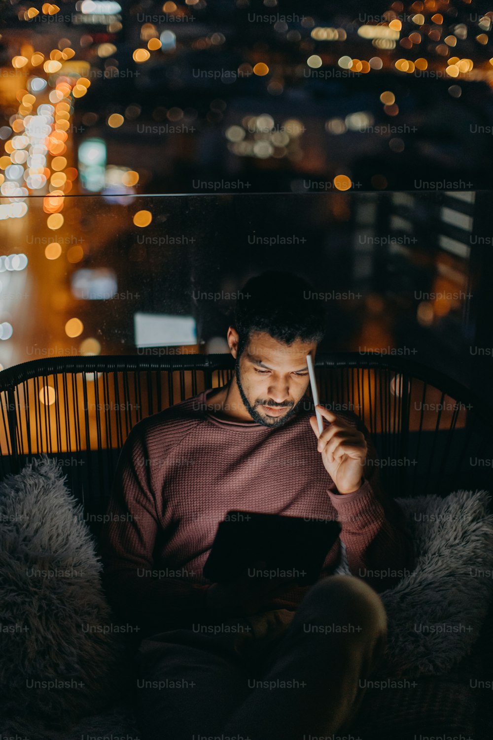 A young African AMerican man sitting on balcony with urban view and using tablet at night