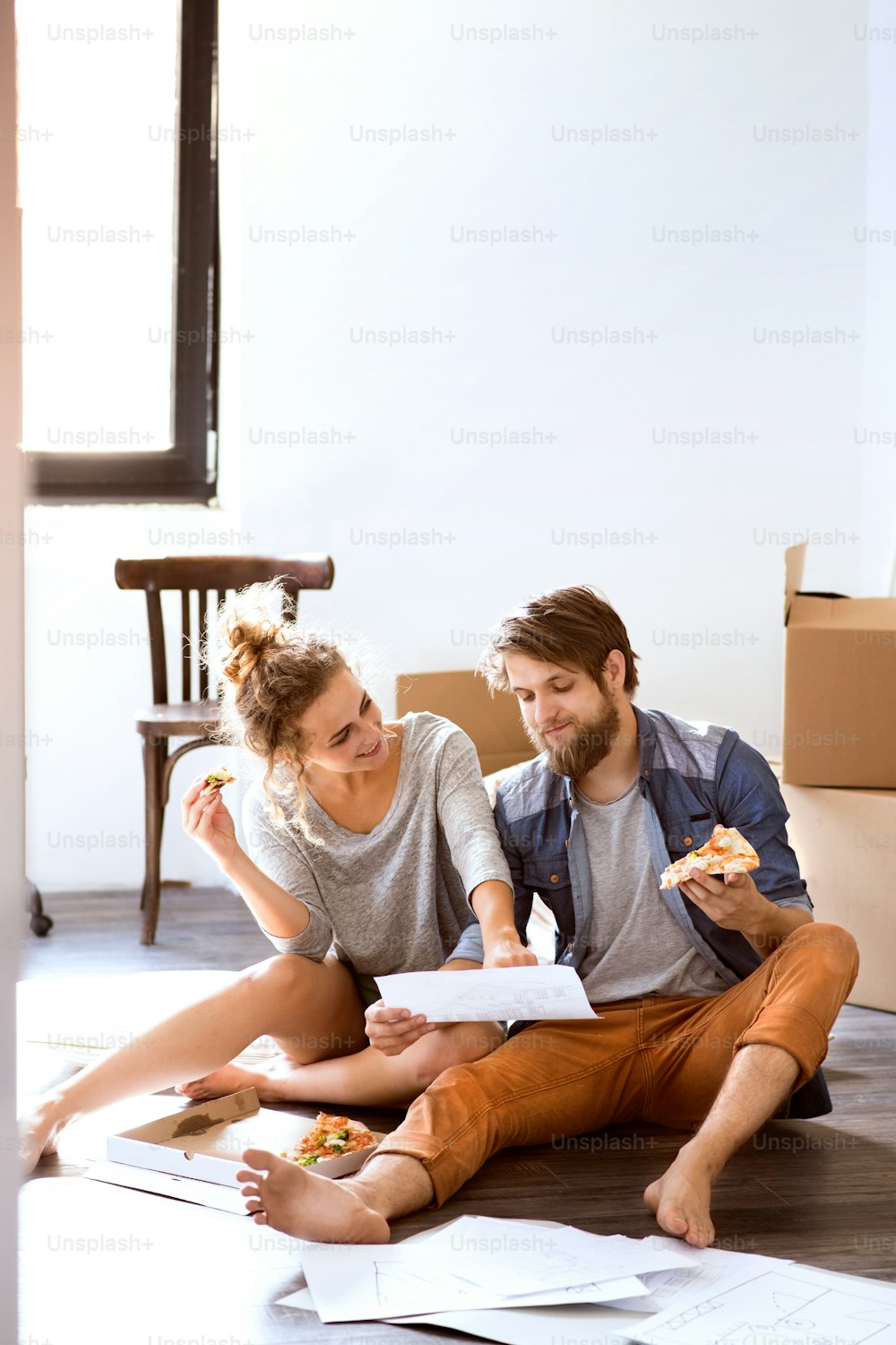 Young married couple moving in new house, sitting on the floor near cardboard boxes, eating pizza, looking at plans.