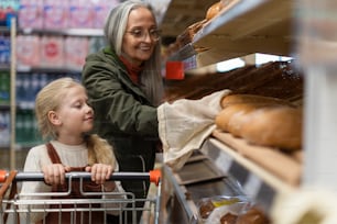 Grandmother with her granddaughter choosing and buying bread in supermarket.