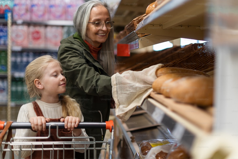 Avó com a neta escolhendo e comprando pão no supermercado.