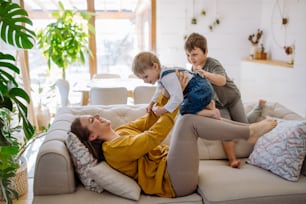 A young cheerful mother playing with her little children and having fun when lifting them up on sofa.