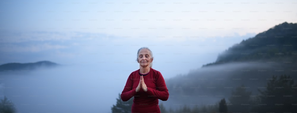 Una mujer mayor haciendo ejercicio de respiración en la naturaleza temprano en la mañana con niebla y montañas en el fondo.