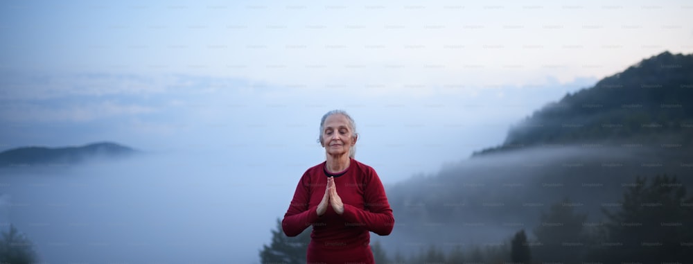 Uma mulher idosa fazendo exercícios de respiração na natureza no início da manhã com neblina e montanhas ao fundo.