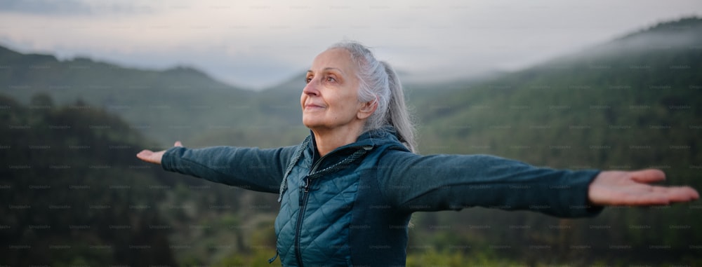Uma mulher idosa fazendo exercícios de respiração na natureza no início da manhã com neblina e montanhas ao fundo.
