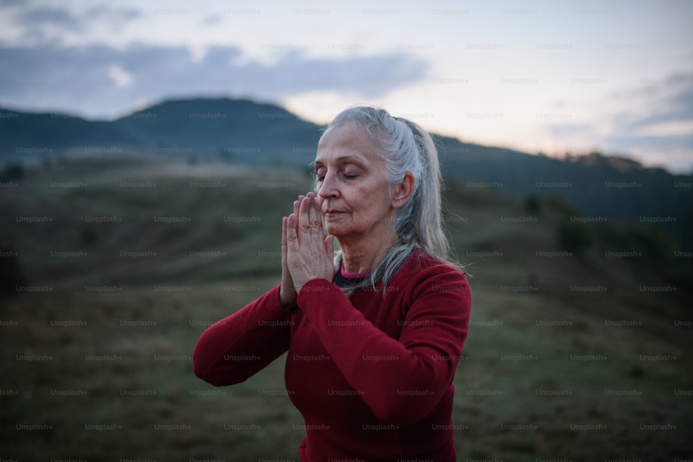 A senior woman doing breathing exercise in nature on early morning with fog and mountains in background.