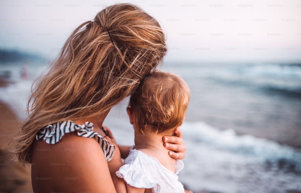 Un primer plano de una madre joven con una niña pequeña en la playa en vacaciones de verano, vista trasera.