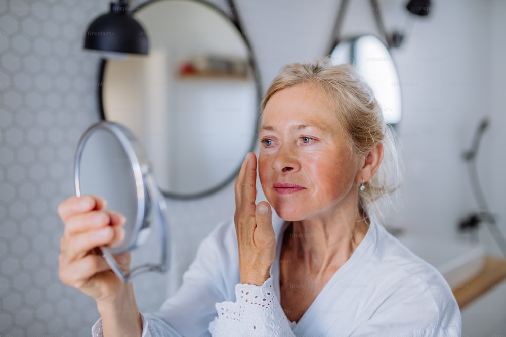 Eine schöne ältere Frau im Bademantel schaut auf Spiegel und trägt natürliche Gesichtscreme im Badezimmer auf, Hautpflegekonzept.