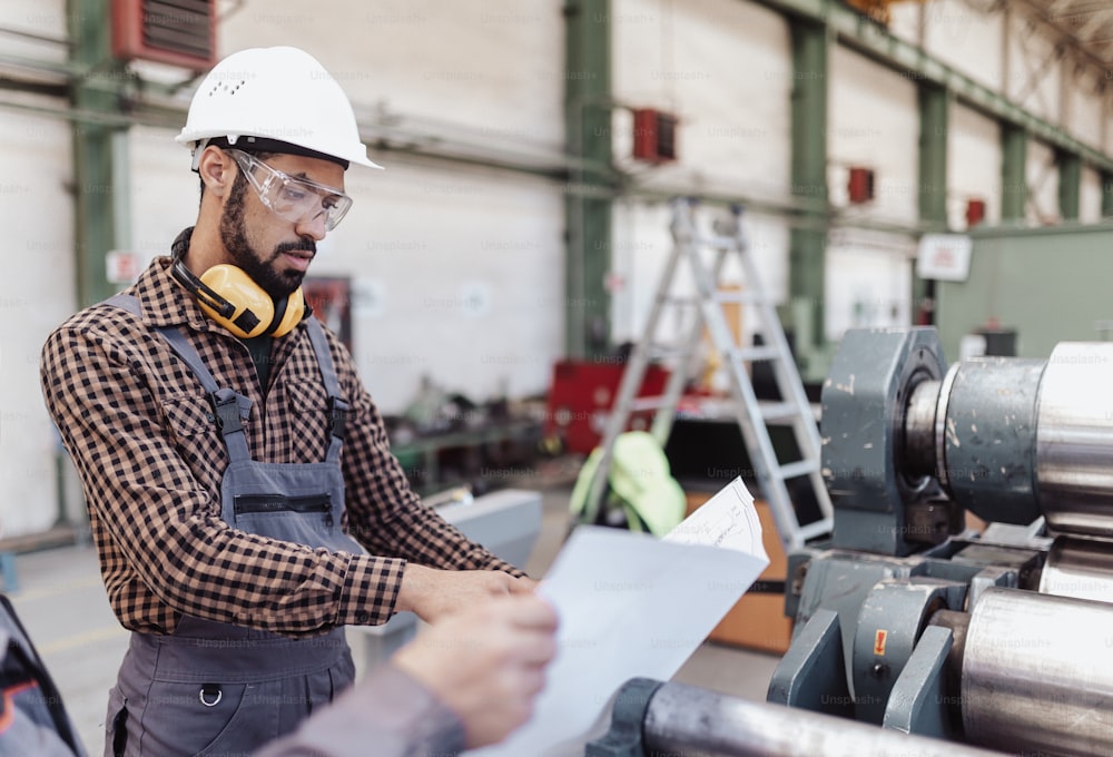 Un trabajador de la industria pesada con auriculares de seguridad y casco en una fábrica industrial sosteniendo planos