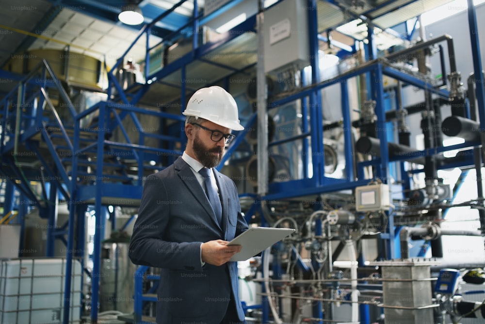 A chief Engineer in the hard hat walks through industrial factory while holding tablet.
