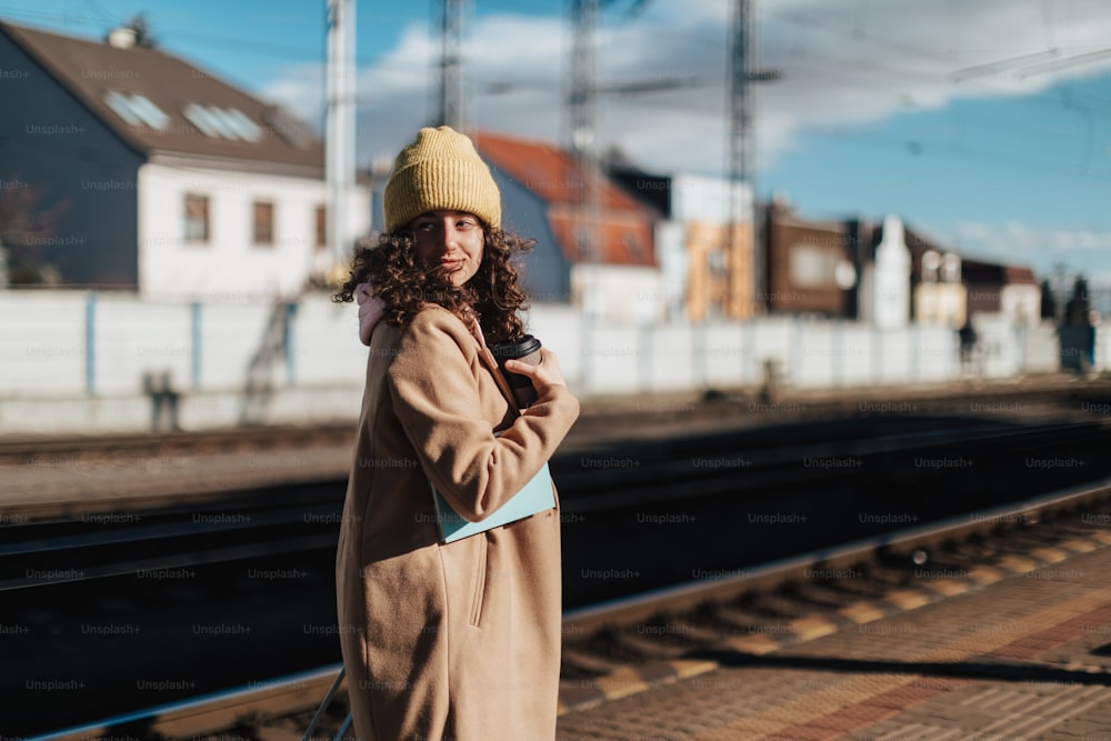Uma mulher jovem viajante feliz com xícara de café na plataforma da estação de trem durante o pôr do sol