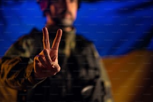 Un soldato in uniforme militare che mostra il segno della pace con la bandiera dell'Ucraina sullo sfondo.