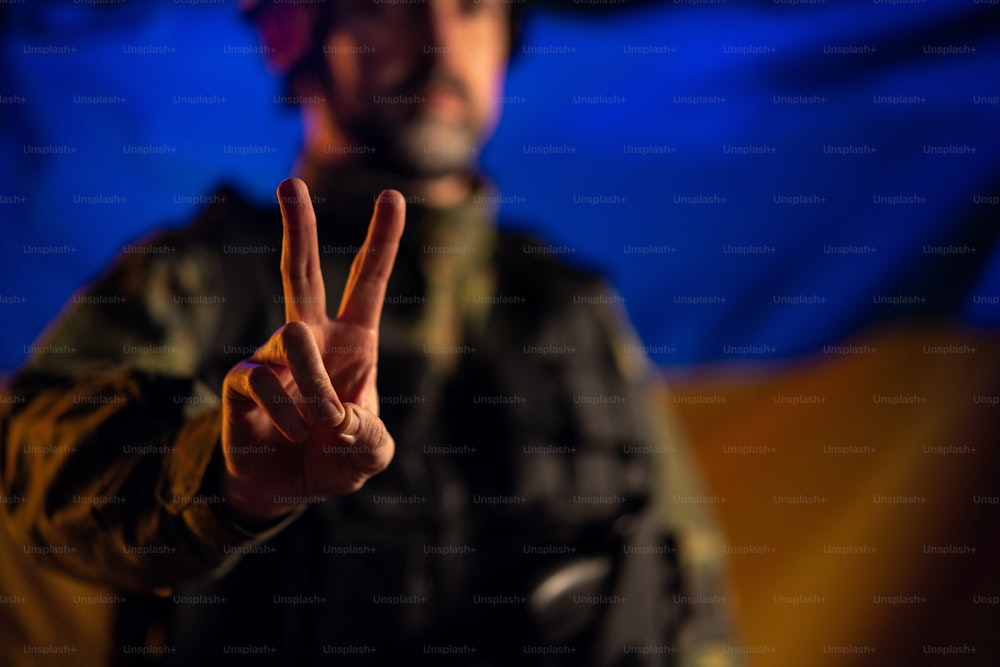 A soldier in military uniform showing peace sign with Ukraine flag in background.