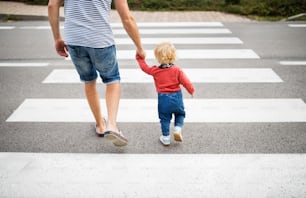 Nicht erkennbarer Vater mit kleinem Jungen, der die Straße auf dem Zebrastreifen überquert. Sommertag. Rückansicht.
