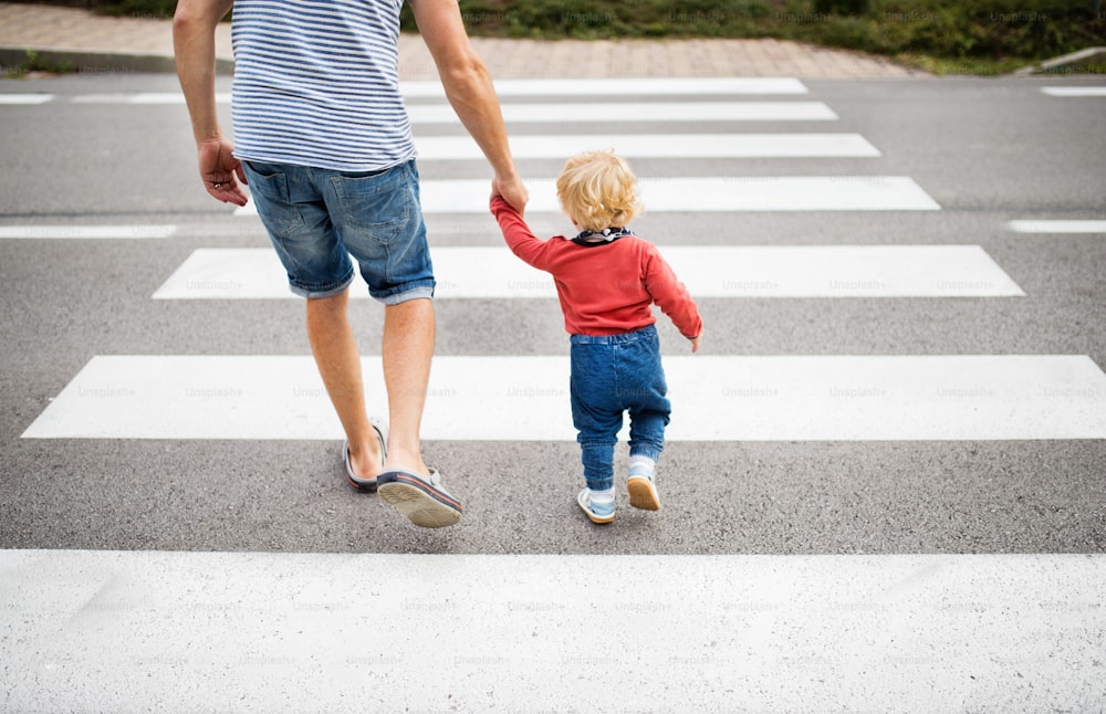 小さな男の子と見分けのつかない父親が横断歩道で道路を横断している。夏の日。背面図。