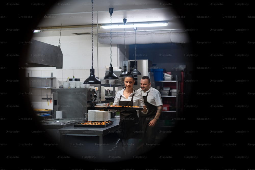 Ein glücklicher Koch und Koch, der in der Restaurantküche an ihren Gerichten arbeitet, erschossen durch ein kreisförmiges Türfenster.