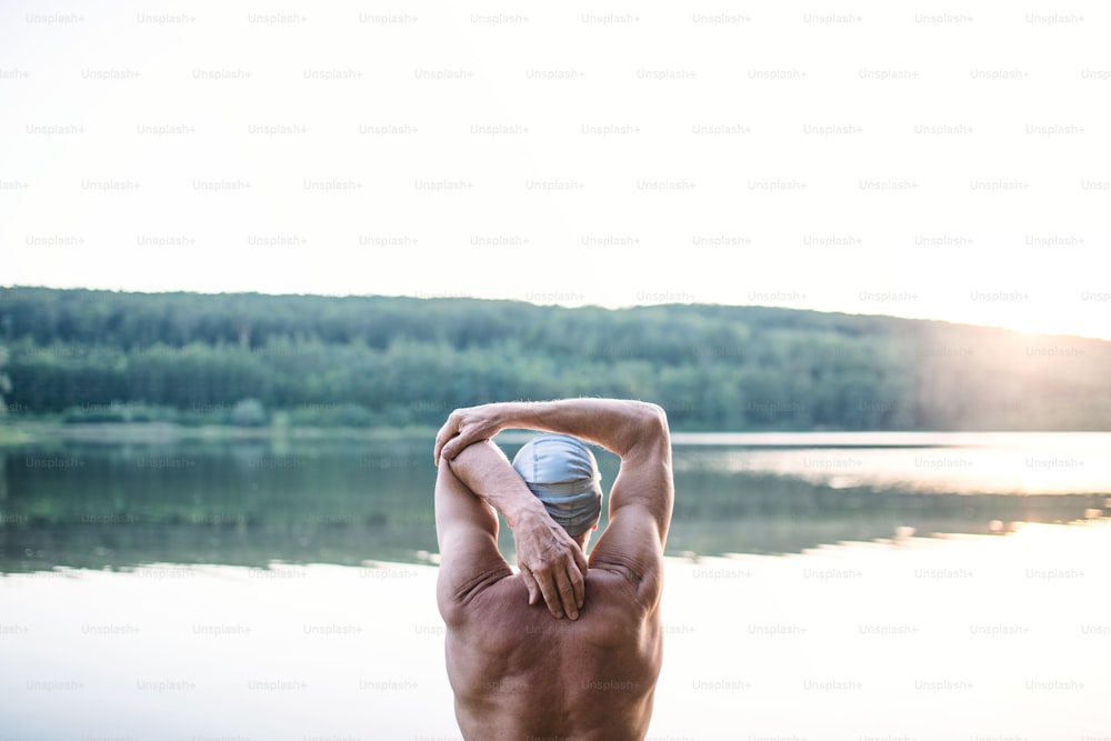 Rear view of senior man standing by lake outdoors before swimming, stretching. Copy space.