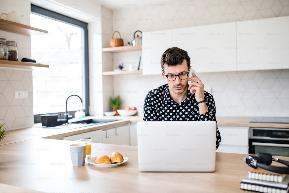 Jeune homme avec ordinateur portable et smartphone assis dans la cuisine, travaillant. Un concept de bureau à domicile.