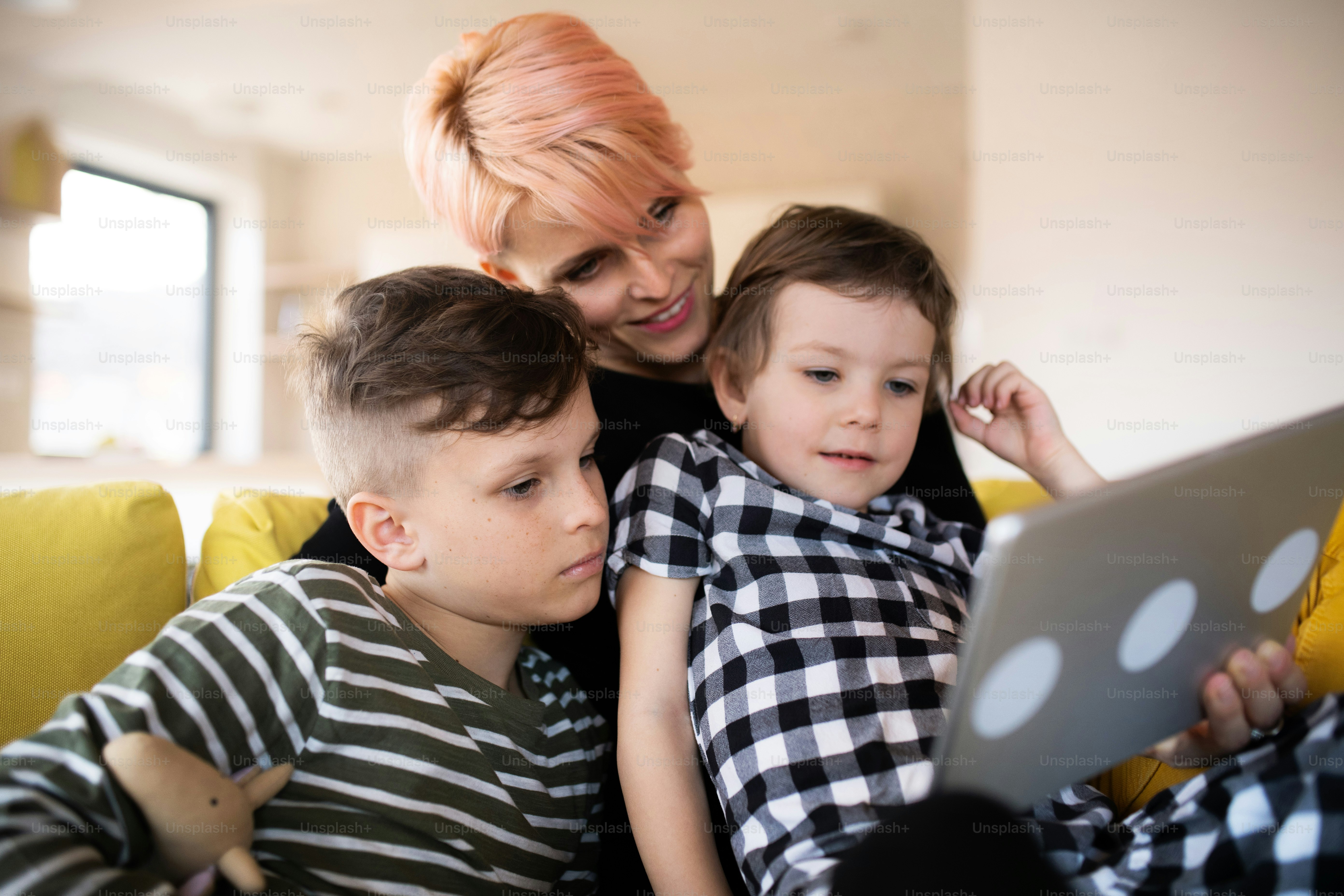 A young unrecognizable woman with two children sitting on sofa indoors at home, using tablet.