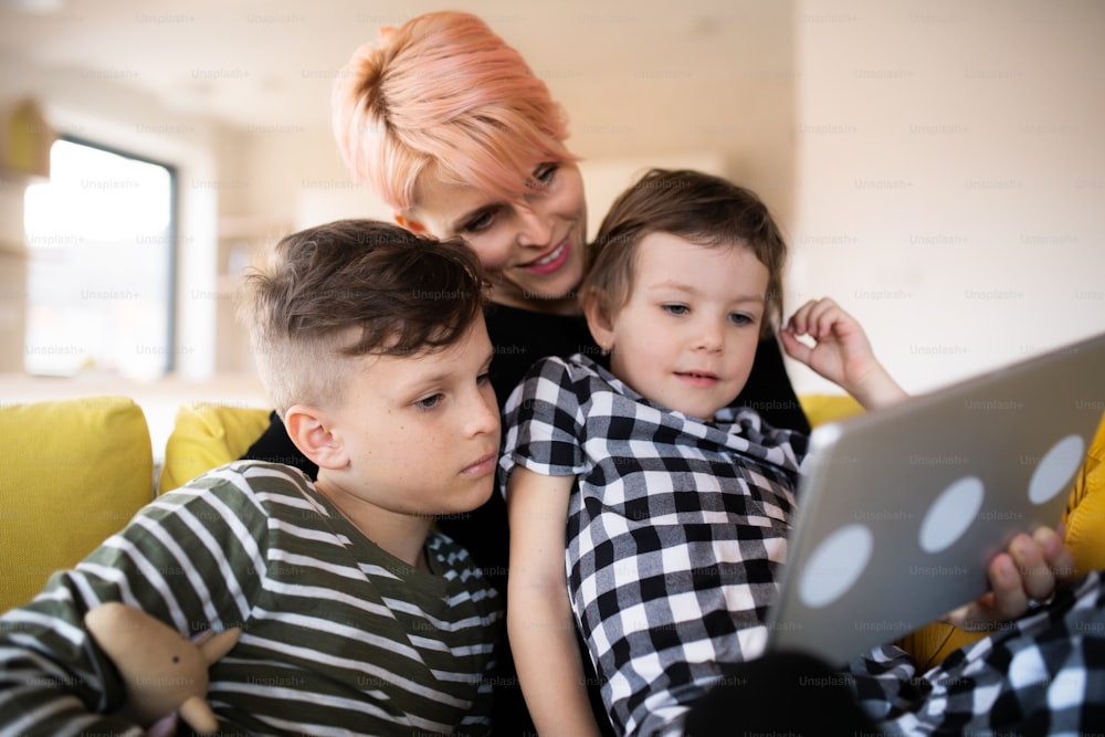 Una giovane donna irriconoscibile con due bambini seduta sul divano all'interno di casa, usando un tablet.