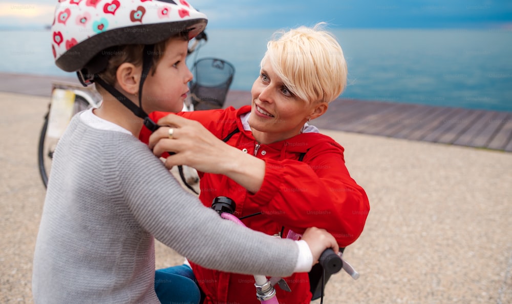 Mother and small daughter with bicycle outdoors on beach, putting on a helmet.