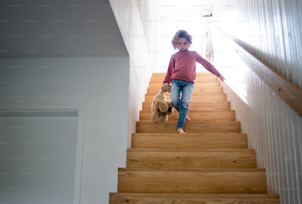 A front view of cute small girl walking down wooden stairs indoors at home.