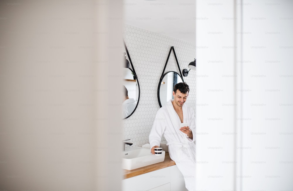 Jeune homme debout dans la salle de bain le matin, utilisant un smartphone.
