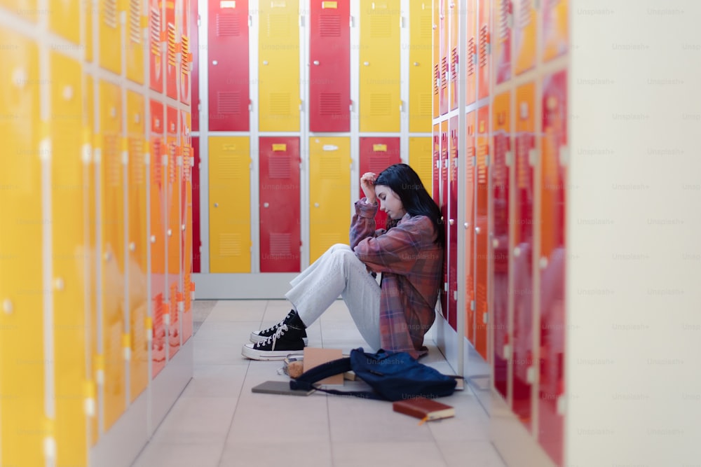 Triste studente adolescente seduto in un corridoio vicino a armadietti colorati e libro da imballaggio nello zaino nel corridoio del campus, concetto di ritorno a scuola.