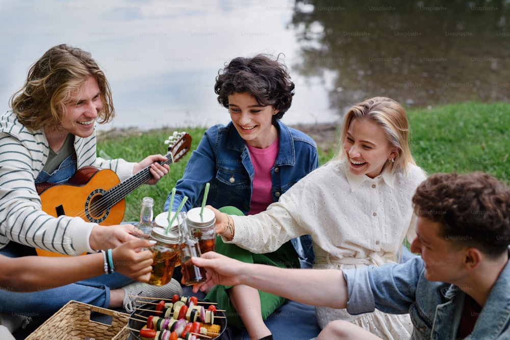 A group of young friends having fun on picnic near a lake, sitting on blanket and toasting with drinks.