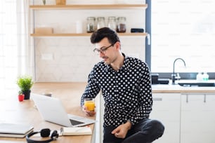Young man with laptop sitting in kitchen, holding juice. A home office concept.