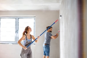 Beautiful young couple having fun and painting walls using paint rollers in their new house. Home makeover and renovation concept.