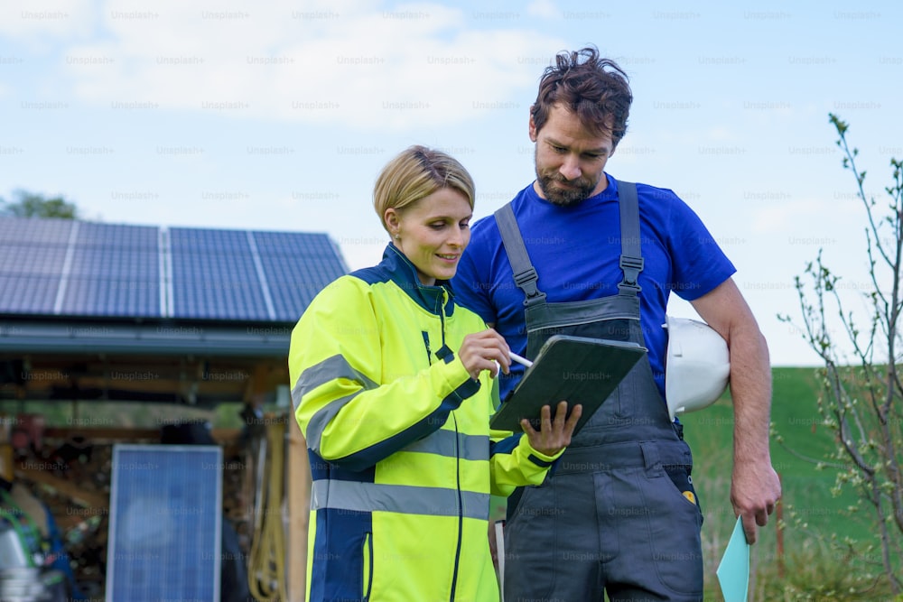 Un hombre y una mujer ingenieros instaladores solares con tableta mientras instalan un sistema de paneles solares en la casa.