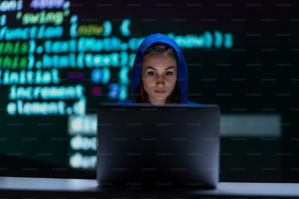 フードをかぶった匿名のハッカーの女性が、夜の暗い部屋でコンピューターを操作し、サイバー戦争のコンセプトを描いています。