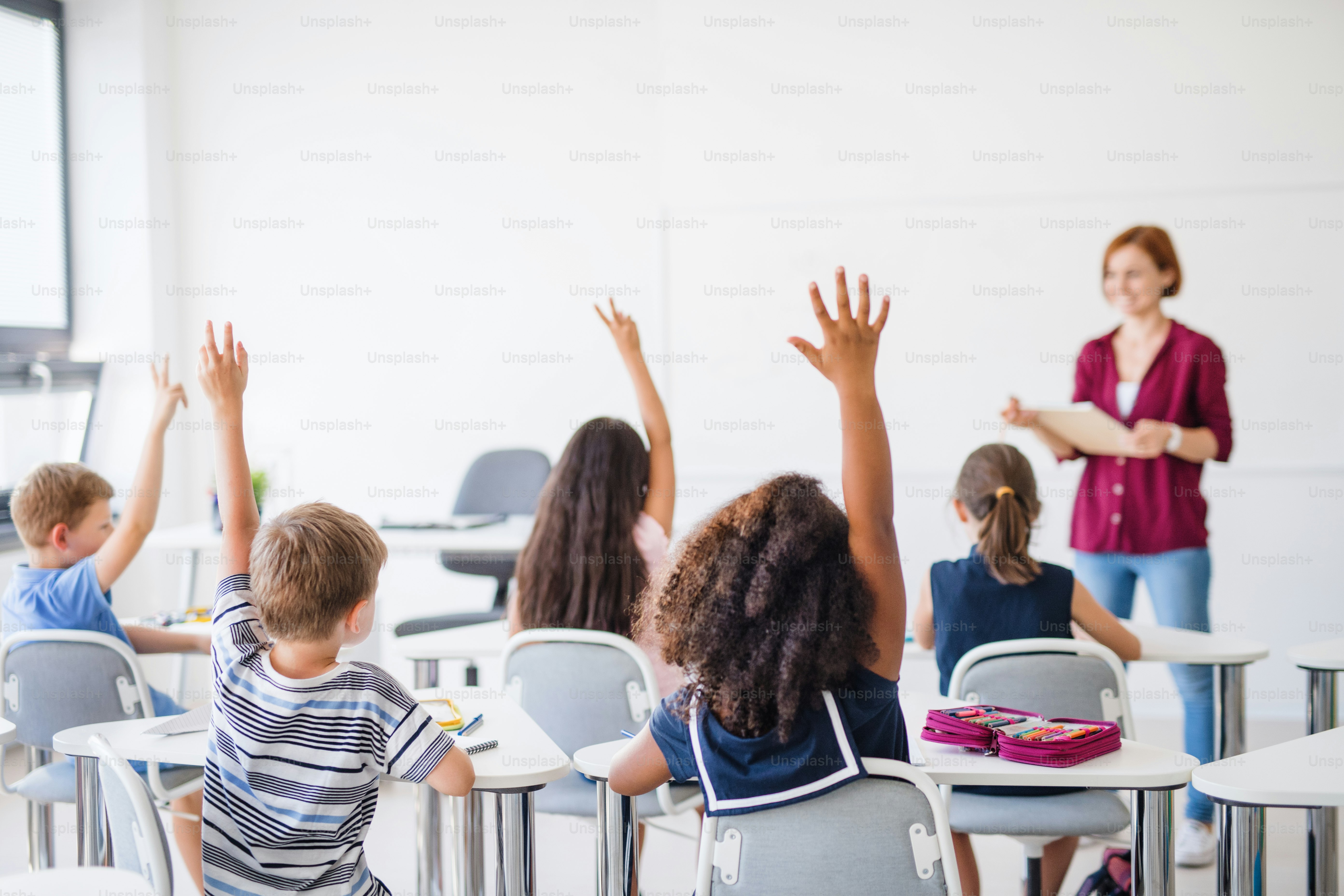 Rear view of school children sitting at the desk in classroom on the lesson, raising hands