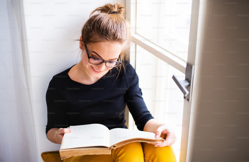 Uma jovem estudante universitária feliz com um livro sentado no peitoril da janela em casa, estudando.