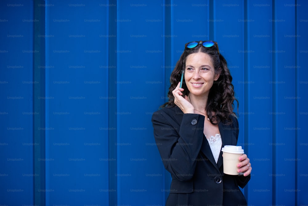 Eine erfolgreiche, glückliche Geschäftsfrau, die morgens in der Stadtstraße pendelt, mit dem Handy telefoniert und einen Kaffeebecher in der Hand hält.