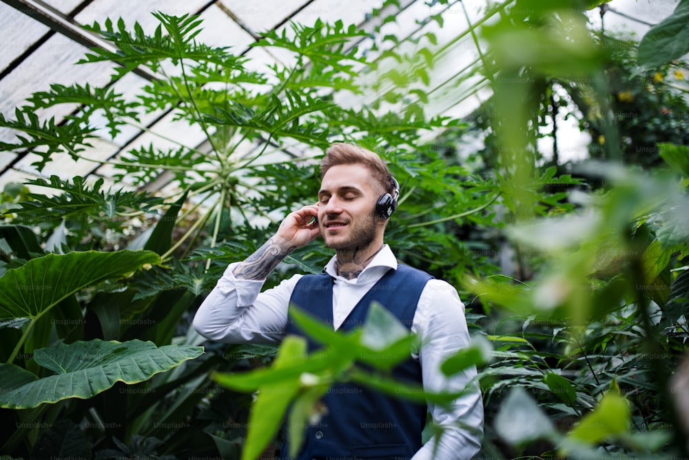 Jeune homme avec des écouteurs debout dans le jardin botanique, écoutant de la musique. Concept d’entreprise verte.