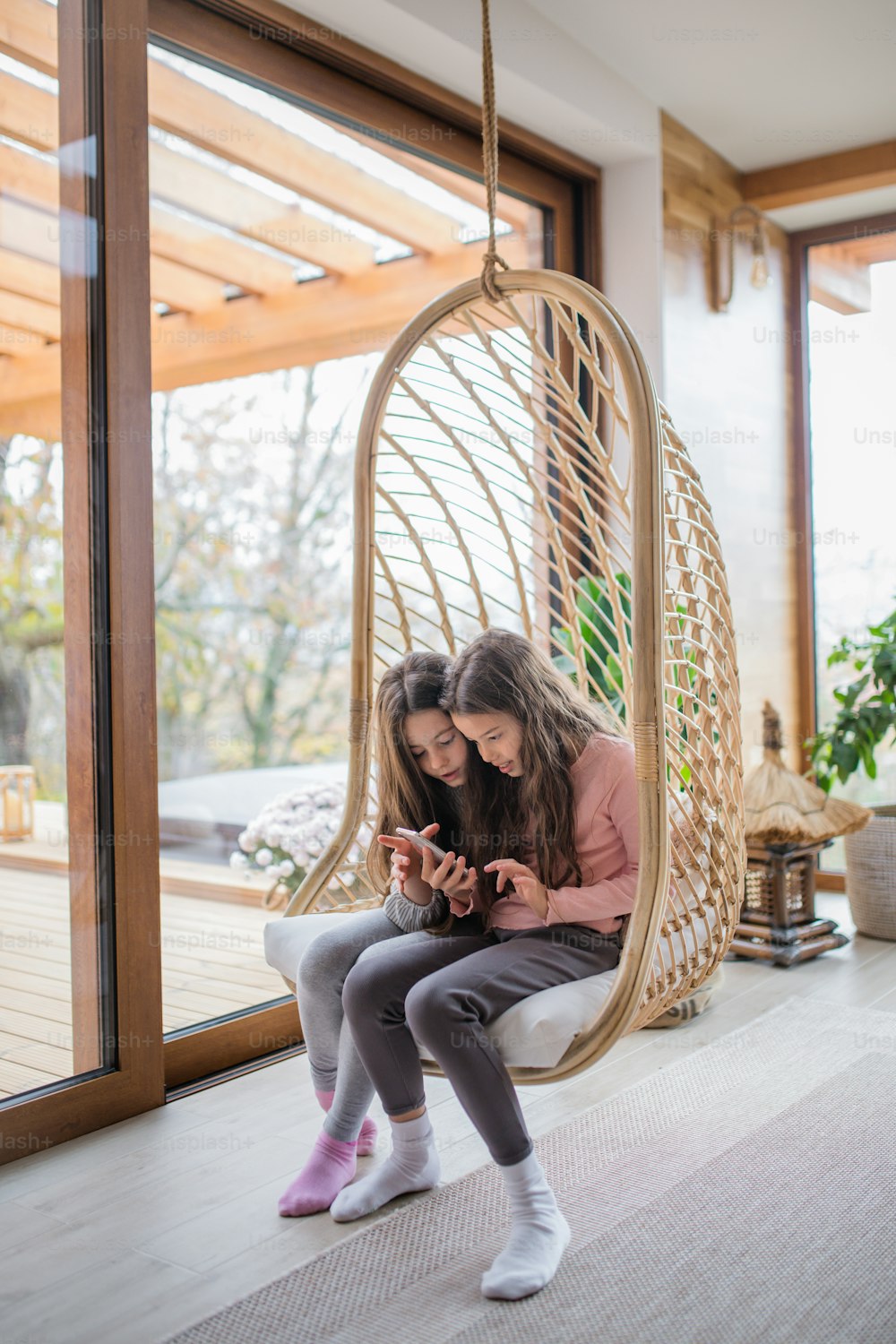 Happy little sisters sitting in a wicker rattan hang chair and using smartphone indoors in conservatory.