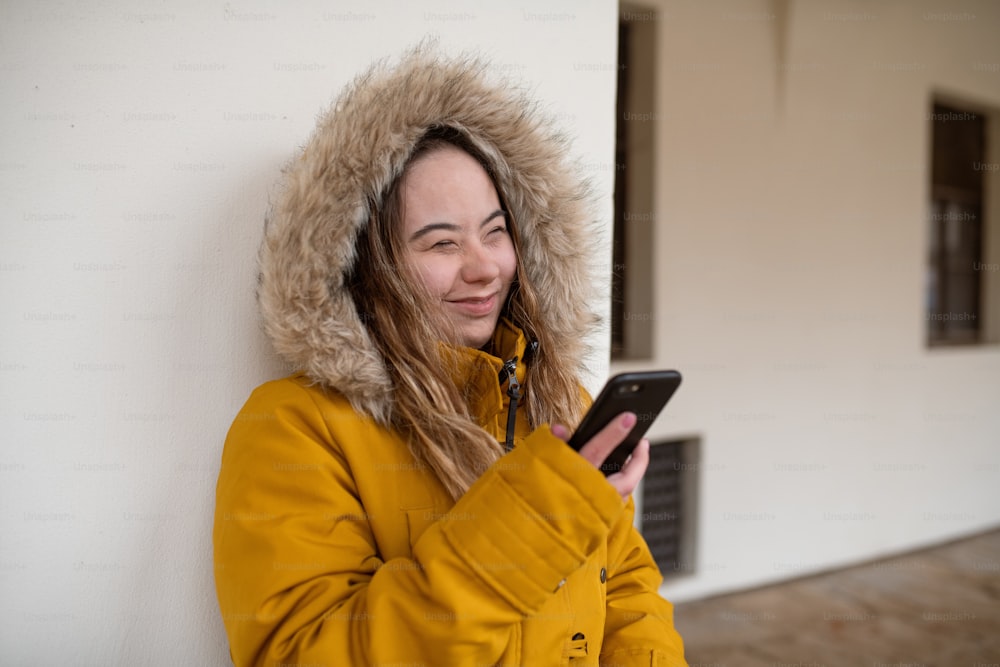A happy young woman with Down syndrome weraing parka, leaning the wall and using smartphone.