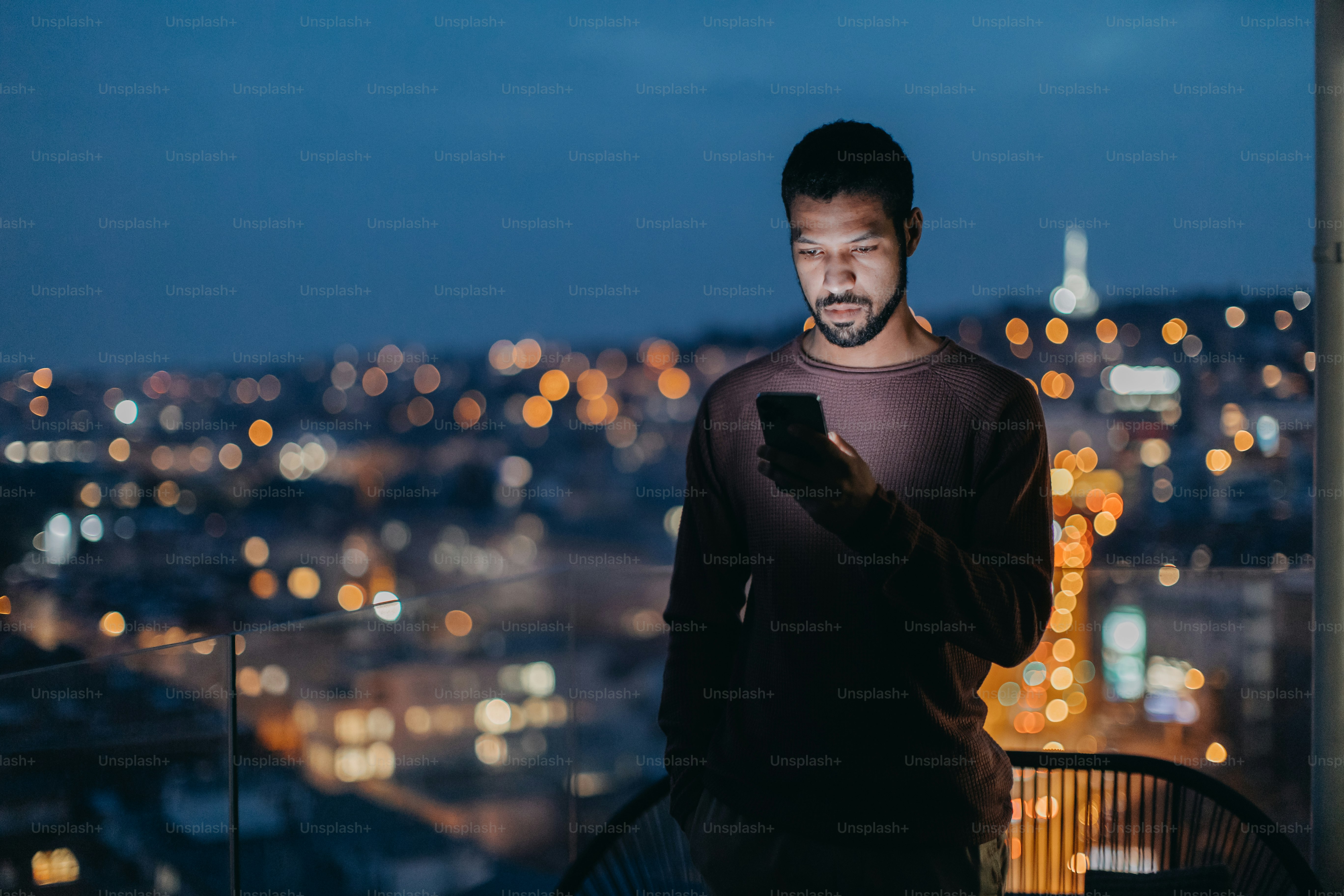 A young man with a smart phone looks sad, standing at balcony with city view in evening at home