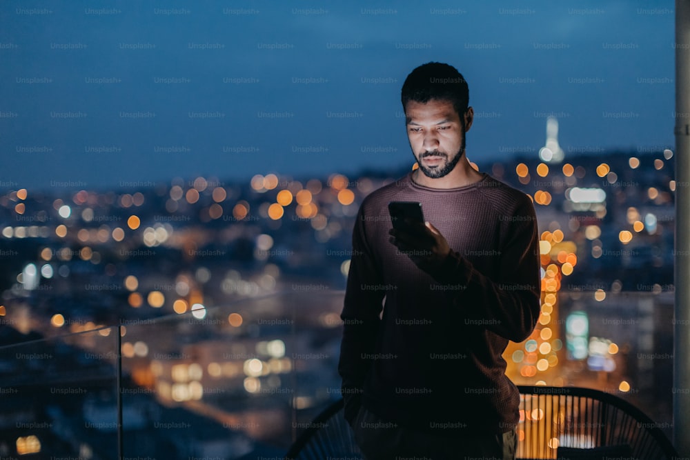 A young man with a smart phone looks sad, standing at balcony with city view in evening at home.