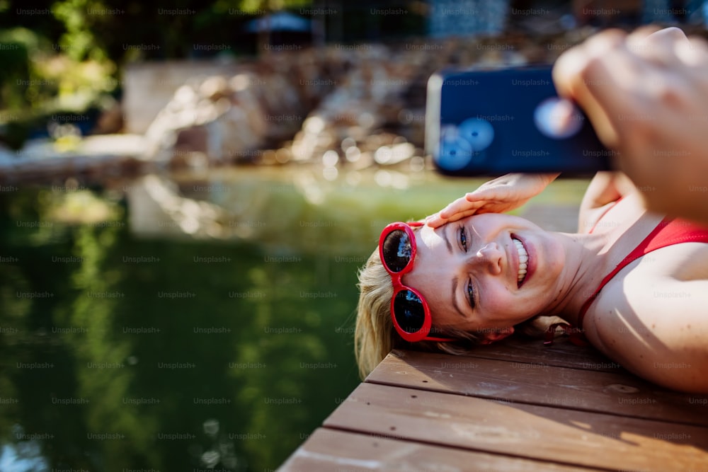 Rlaxed woman relaxing on the pier, lying and taking selfie near natural lake in summer.