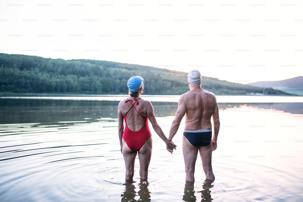 Rear view of senior couple in swimsuit standing by lake outdoors before swimming.