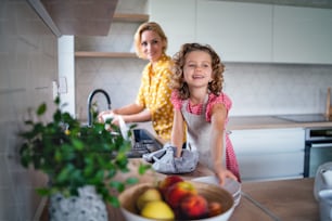 Uma menina pequena bonita com a mãe dentro de casa na cozinha de casa, lavando os pratos.