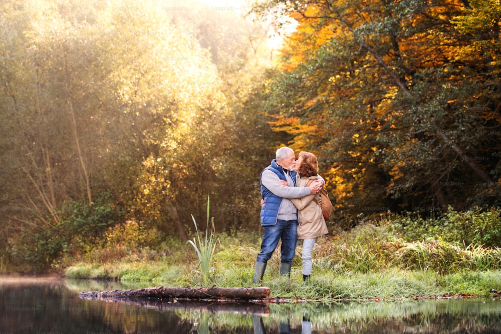 Active senior couple on a walk in a beautiful autumn nature. A woman and man by the lake in the early morning, kissing.