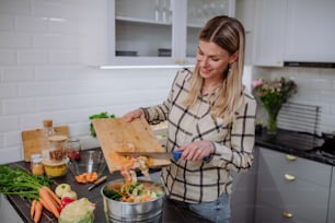 Une femme jetant des boutures de légumes dans un seau à compost dans la cuisine.