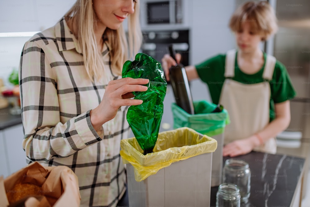 Una madre con su hija tirando botellas vacías de plástico y vidrio en un contenedor de reciclaje en la cocina.